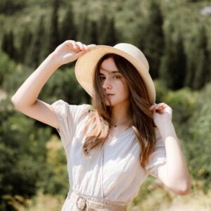 Young woman posing outdoors in a white dress and sun hat under bright sunlight.